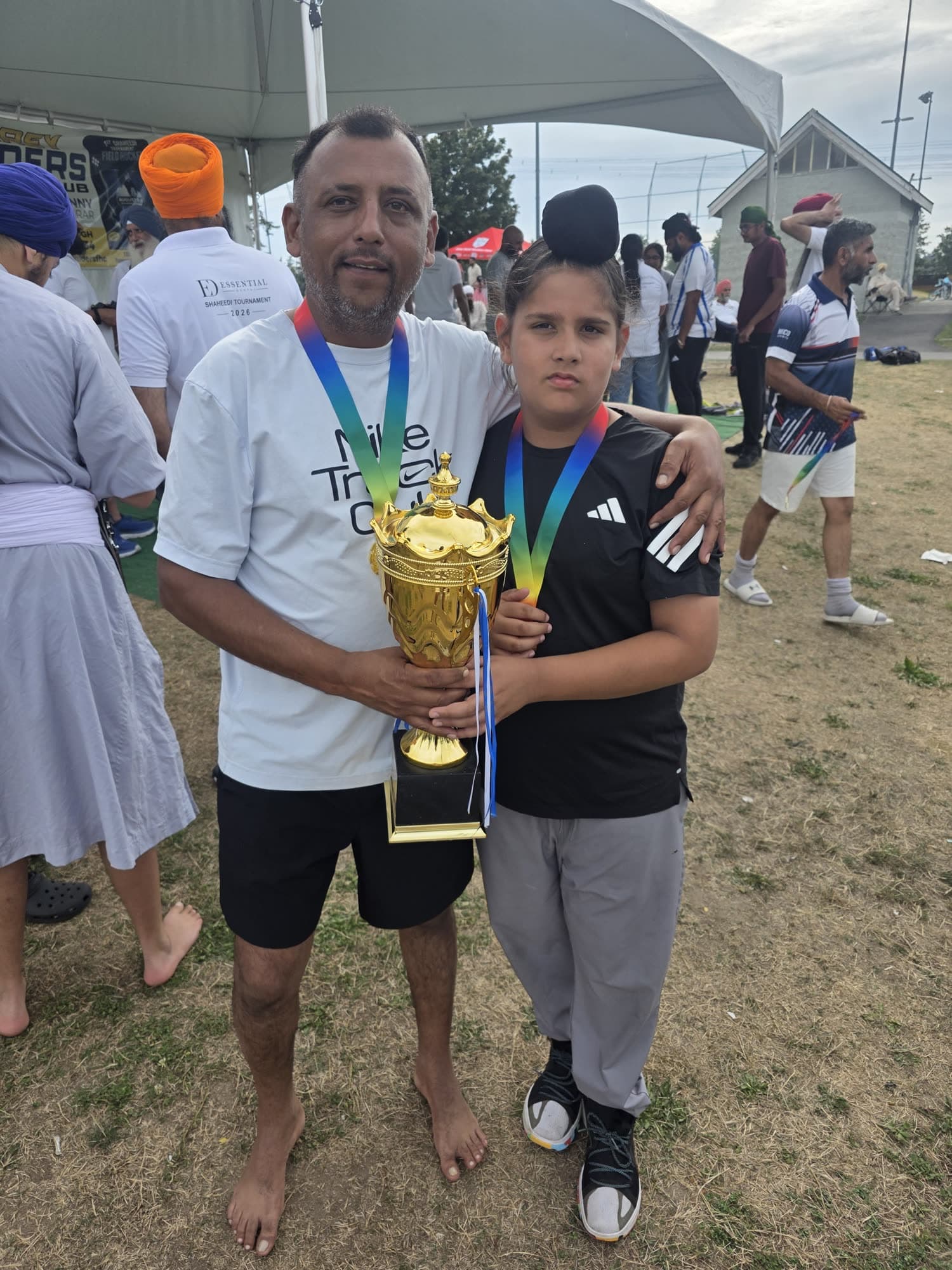 Mr. Johal holding the gold trophy with his arm around his son Avitaj, both wearing tournament medals, under the Surrey Raiders tent